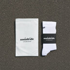 Ein Paar weisse Radsocken mit einem schwarzen "Socialride Cycling Socks"-Etikett, flach auf grauem Asphalt liegend, daneben ein weisser Umschlag mit demselben Branding.