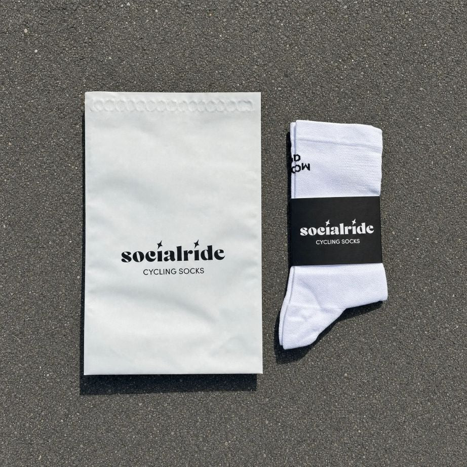 Ein Paar weisse Radsocken mit einem schwarzen "Socialride Cycling Socks"-Etikett, flach auf grauem Asphalt liegend, daneben ein weisser Umschlag mit demselben Branding.