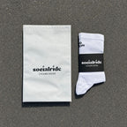 Ein Paar weisse Radsocken mit einem schwarzen "Socialride Cycling Socks"-Etikett, flach auf grauem Asphalt liegend, daneben ein weisser Umschlag mit demselben Branding.