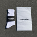 Ein Paar weisse Radsocken mit einem schwarzen "Socialride Cycling Socks"-Etikett, flach auf grauem Asphalt liegend, daneben ein weisser Umschlag mit demselben Branding.